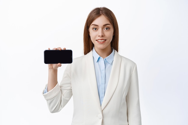 プロの服を着た企業の女性は、空の携帯電話の画面を水平に表示し、アプリや会社のサイトを示し、白い壁に立っています
