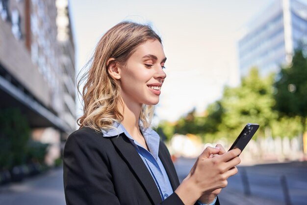 Corporate woman stands on street and texts message on mobile phone smiling while looking at smartpho