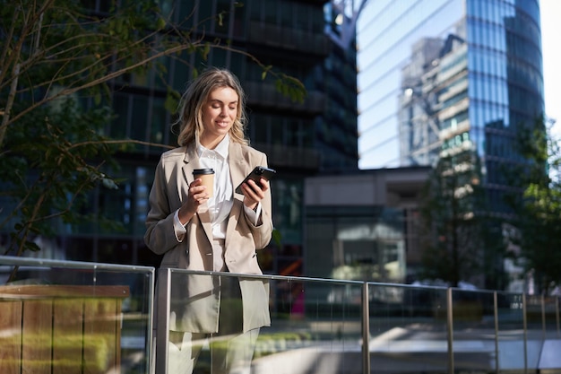 La donna corporativa in tuta beve il caffè tiene il telefono cellulare in mano si trova da sola sulla strada della città