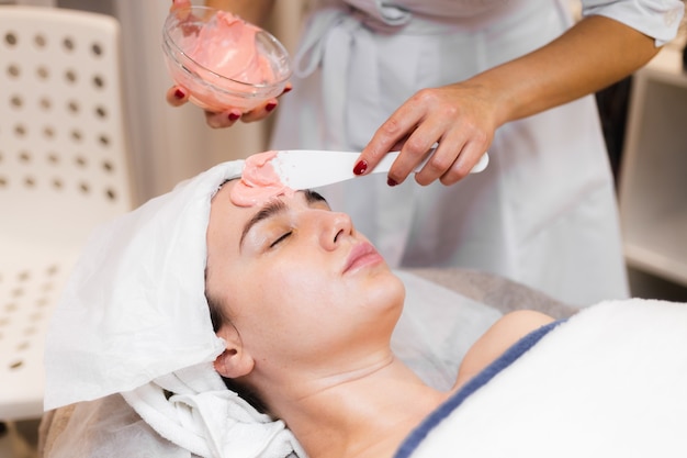 美容師は、女性の顔にヘラでアルギン酸塩マスクを適用します。