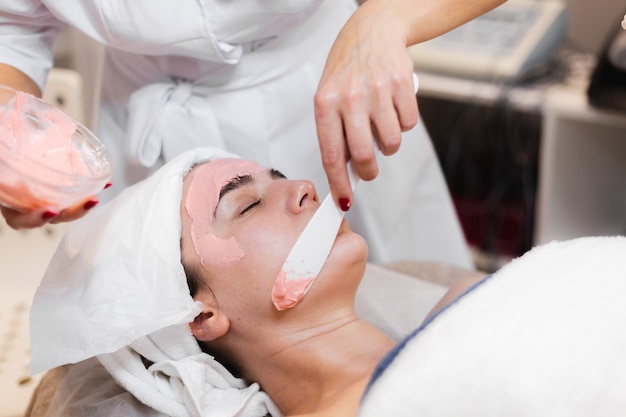 Cosmetologist applies alginate mask with spatula on face of woman.