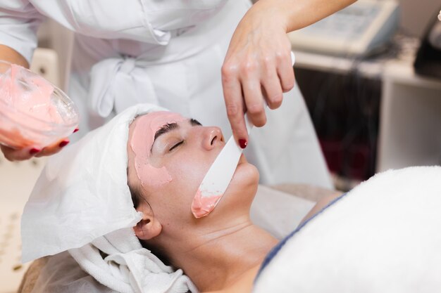 美容師は、女性の顔にヘラでアルギン酸塩マスクを適用します。