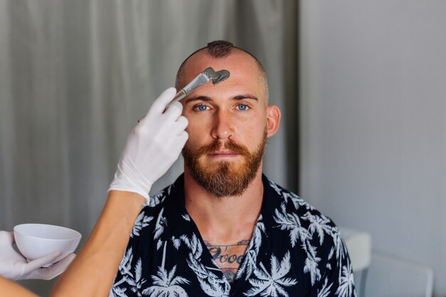 Cosmetologist apply clay mask with brush on young handsome mans face in beauty clinic