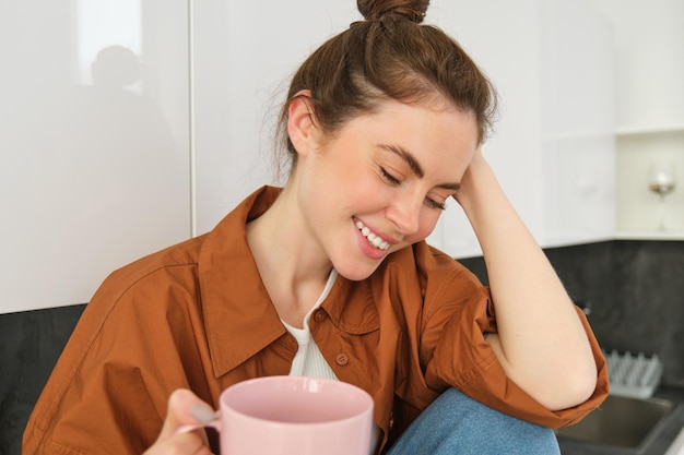 無料写真 居心地の良い瞬間 ⁇ 幸せな若い女性がキッチンに座ってコーヒーを飲み ⁇ 彼女の顔には優しい笑顔があります ⁇