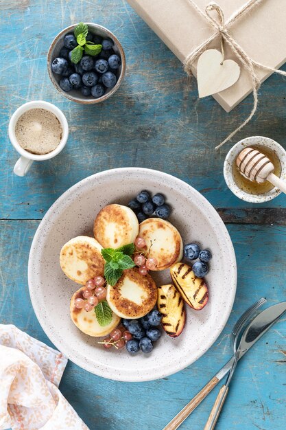코티지 치즈 팬케이크 치즈케이크 리코타 프리터와 신선한 블루베리 건포도, 복숭아를 접시에 올려 휴일을 위한 건강하고 맛있는 아침 식사 푸른 나무 배경