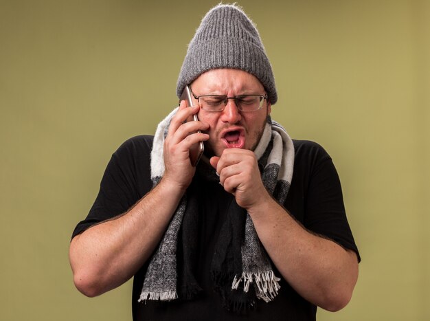 Coughing middle-aged ill male wearing winter hat and scarf speaks on phone isolated on olive green wall