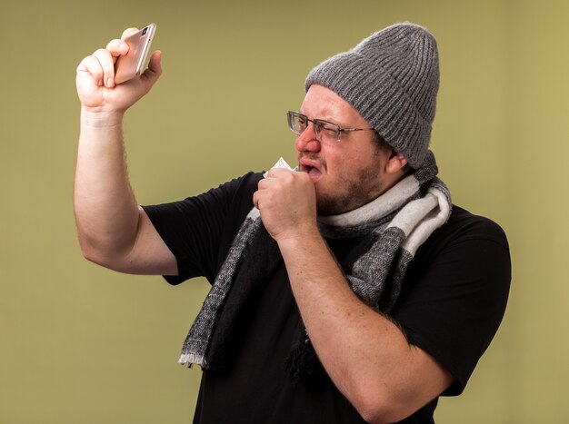 Coughing middle-aged ill male wearing winter hat and scarf take a selfie isolated on olive green wall