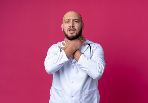 Coughing young bald male doctor wearing medical robe and stethoscope grabbed throat isolated on pink wall