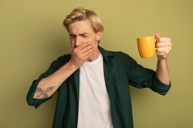 お茶のカップと手で覆われた口を保持している緑のTシャツを着て咳をする若いブロンドの男