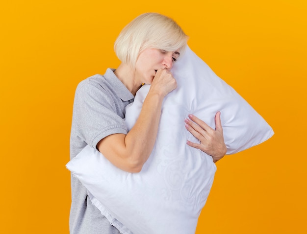 Coughing young blonde ill woman hugs pillow isolated on orange wall