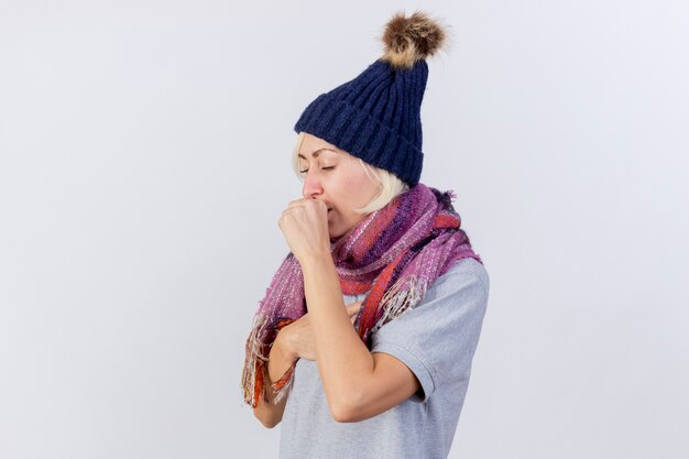 Coughing young blonde ill woman wearing winter hat and scarf holds fist close to mouth isolated on white wall