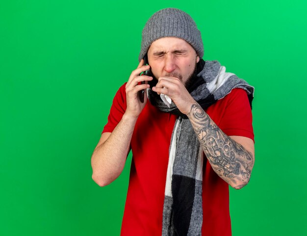 冬の帽子とスカーフを身に着けている若い白人の病気の男性の咳は、電話で話しているティッシュを保持します