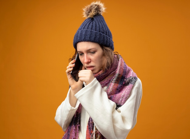 白いローブとスカーフと冬の帽子を身に着けて見下ろしている咳の若い病気の女の子は、オレンジ色に分離された口に手を置いて電話で話します