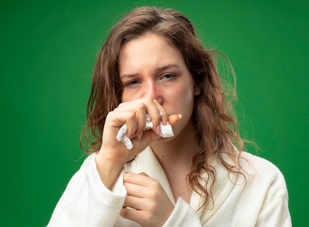 Coughing young ill girl looking straight ahead wearing white robe holding hand on mouth isolated on green