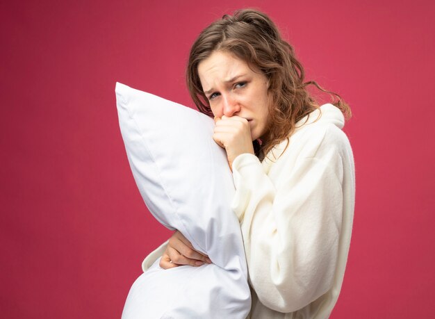 Coughing young ill girl looking straight ahead wearing white robe hugged pillow isolated on pink