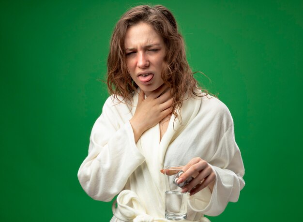 Coughing young ill girl wearing white robe holding glass of water grabbed throat isolated on green