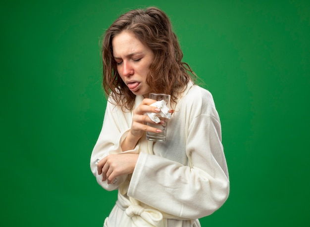 Coughing young ill girl with closed eyes wearing white robe holding glass of water isolated on green