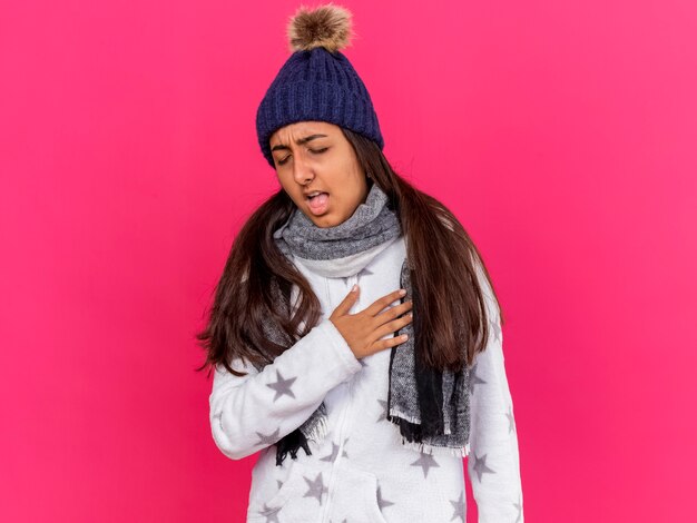Кашель молодая больная девушка с закрытыми глазами в зимней шапке с шарфом, положив руку на сердце, изолированное на розовом фоне