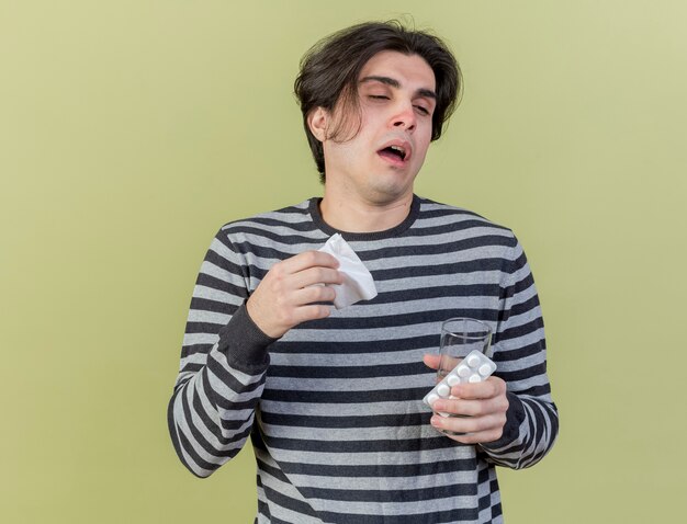 coughing young ill man holding napkin and glass of water with pills isolated on olive green