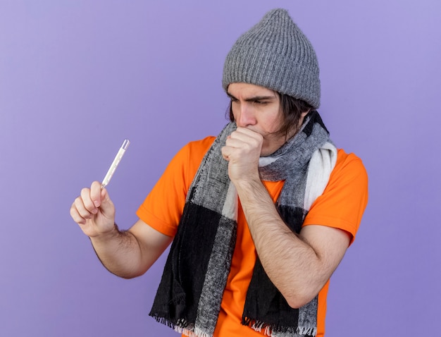 Бесплатное фото Кашель молодой больной человек в зимней шапке с шарфом, держащий и смотрящий на термометр, изолированный на фиолетовом фоне