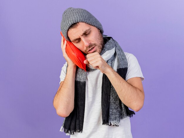 coughing young ill man wearing winter hat with scarf holding hot-water bottle on cheek isolated on purple