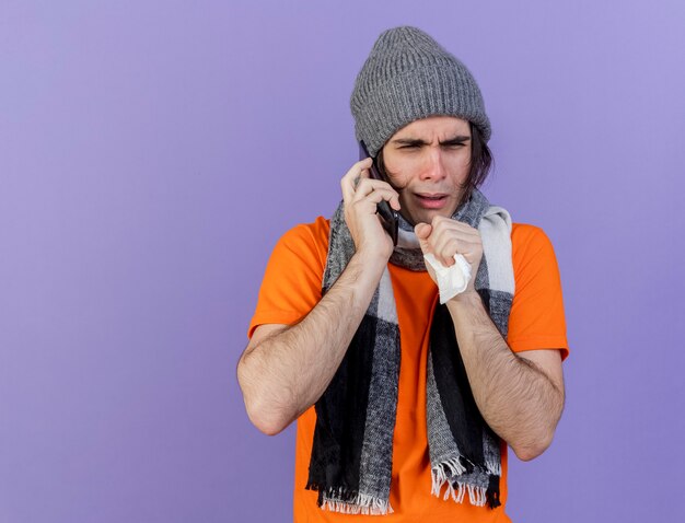 Coughing young ill man wearing winter hat with scarf speaks on phone isolated on purple background