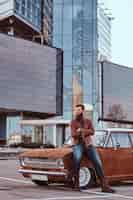 Free photo country style concept. bearded male dressed in brown leather jacket and boots leaning on tuned retro car in the city parking near skyscraper.