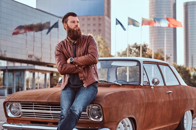 country style concept. bearded male dressed in brown leather jacket and boots leaning on tuned retro car in the city parking near skyscraper.