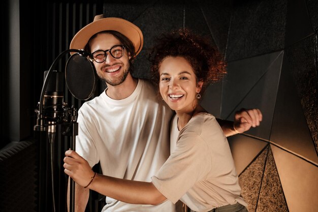 Couple of attractive stylish musicians happily looking in camera working together on new music album in modern sound recording studio