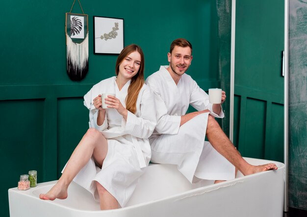 Couple in bathrobes holding cups and posing in bathtub