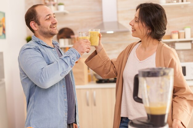 キッチンでスムージーグラスをチリンと鳴らすカップル。陽気な男と女。健康的なのんきで陽気なライフスタイル、食事を食べ、居心地の良い晴れた朝に朝食を準備する