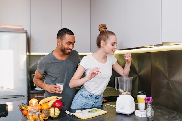 Couple cooking together in the cozy kitchen. Girl puts fruits into a blender, blonde loves a healthy diet. Pair spend time in the modern house.
