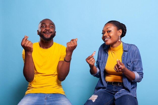 Couple doing money gesture and demanding cash reward while they sit in studio. Man and woman rubbing fingers to ask for payment, feeling optimistic. People using body language to receive income.