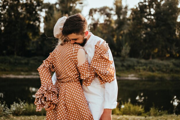 Couple embraces against lake in park Man in white shirt and woman in brown dress have nice summer day