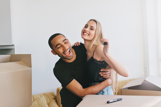 Couple exited about moving in new home, new life. Girl holding keys of new house,  boxes around, man and woman hugging