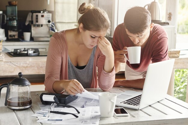 Couple facing financial problem, failing to pay loan in bank. Stressed woman managing family budget, making calculations using laptop and calculator, her husband standing next to her with cup of tea