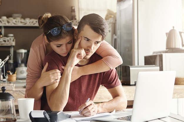 Couple facing financial stress. Young thoughtful bearded man holding index finger on his temple planning family budget at home, using laptop and calculator. His supportive wife hugging him from behind