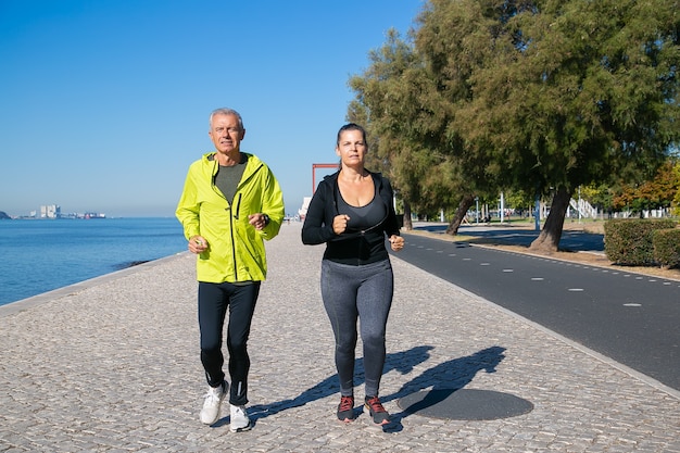 Coppia di appassionati di jogging maturi concentrati che corre lungo la riva del fiume. uomo dai capelli grigi e donna che indossa abiti sportivi, correndo all'esterno. attività e concetto di pensionamento