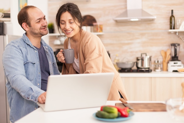 웃는 부엌에서 노트북 앞의 커플. 커피 컵과 아내입니다. 프리랜서 남자와 여자. 현대적인 Wi-Fi 무선 인터넷 기술을 사용하여 집에서 사랑하는 부부의 행복한 사랑의 쾌활한 낭만적인