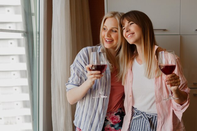 Couple of happy carefree women looking at window and holding glass of wine. Cozy home atmosphere.