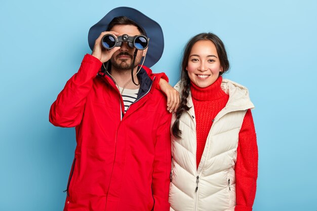 Couple have journey in mountains, look through binoculars, observe landscape, being full of energy, dressed in casual outfits, stand closely, isolated on blue