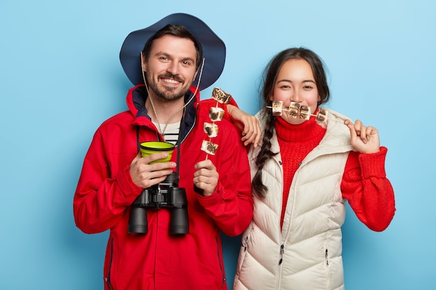 Free photo couple have picnic together, enjoys being alone with nature, eat roasted marshmallow and drink coffee, being in good mood, pose against blue wall