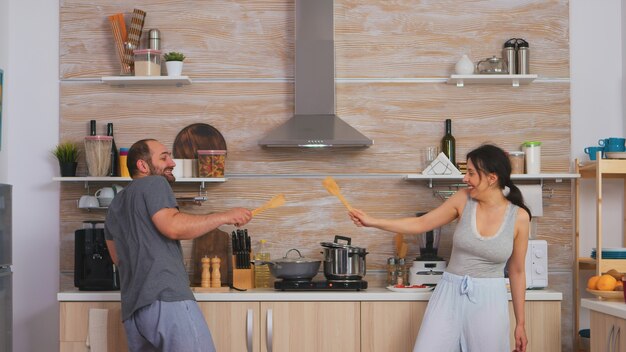 Couple having fun in the kitchen fencing with big spoons during breakfast wearing pajamas. Cheerful carefree joyful funny lovers, fighting wooden spoon, bonding game fight swordplay, happy lifestyle