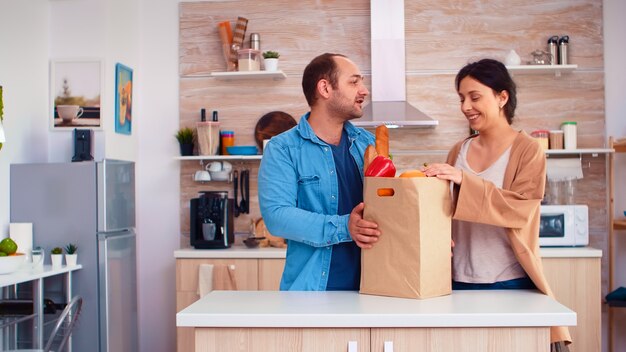 Couple holding paper bag with grocery from supermarket in kitchen. Cheerful happy family healthy lifestyle, fresh vegetables and groceries. Supermarket products shopping lifestyle