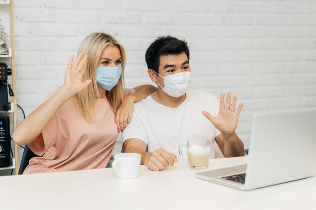 Couple at home with medical masks during the pandemic waving at laptop