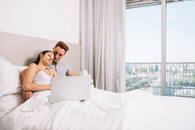 Free photo couple hugging in white bed with laptop