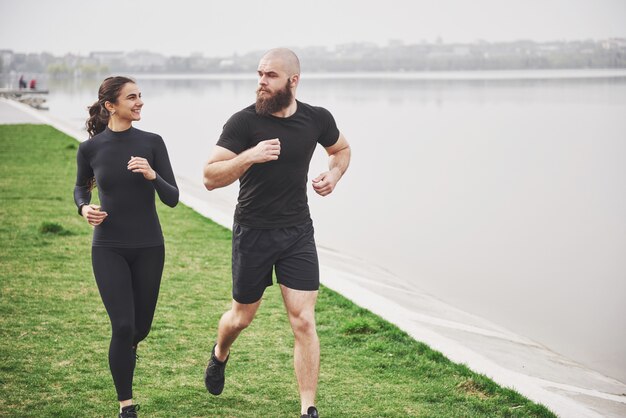 Couple jogging and running outdoors in park near the water. Young bearded man and woman exercising together in morning