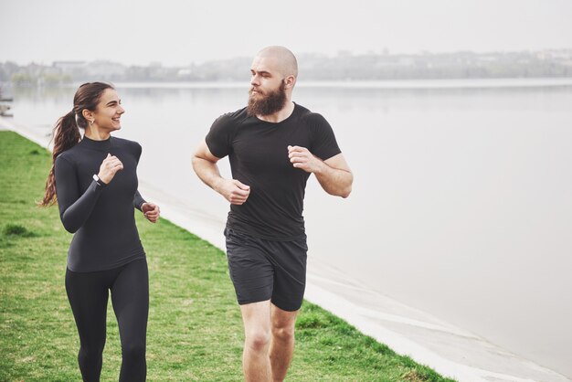 Couple jogging and running outdoors in park near the water. Young bearded man and woman exercising together in morning