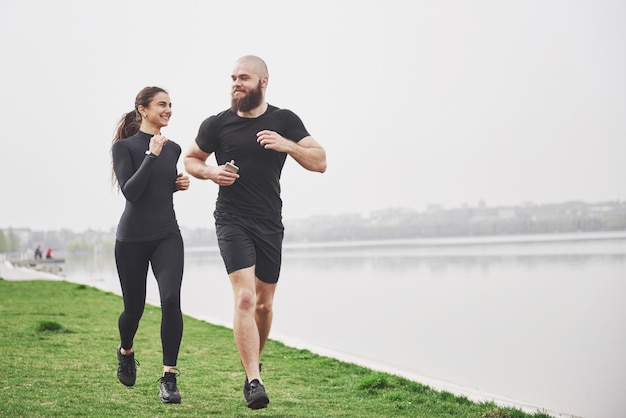 ジョギングや屋外の水の近くの公園で走っているカップル。若いあごひげを生やした男性と女性が朝一緒に運動