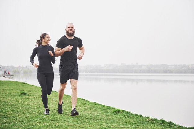 Couple jogging and running outdoors in park near the water. Young bearded man and woman exercising together in morning